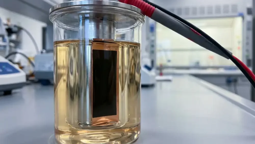 Cylindrical battery electrode submerged in electrolyte inside a glass electrochemical test cell with copper current collector and multimeter probe.