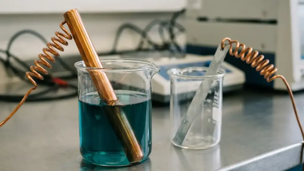 Copper and zinc electrodes in teal electrolyte solution inside laboratory beakers, connected by coiled copper wire on a steel bench.
