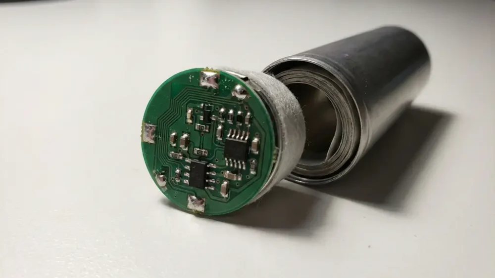 Disassembled lithium-ion cylindrical cell with exposed protection circuit board showing copper traces and miniature components on a lab bench.