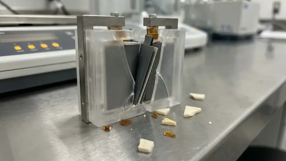 Cracked solid-state battery test cell with warped electrodes and scattered ceramic electrolyte fragments on a laboratory bench.