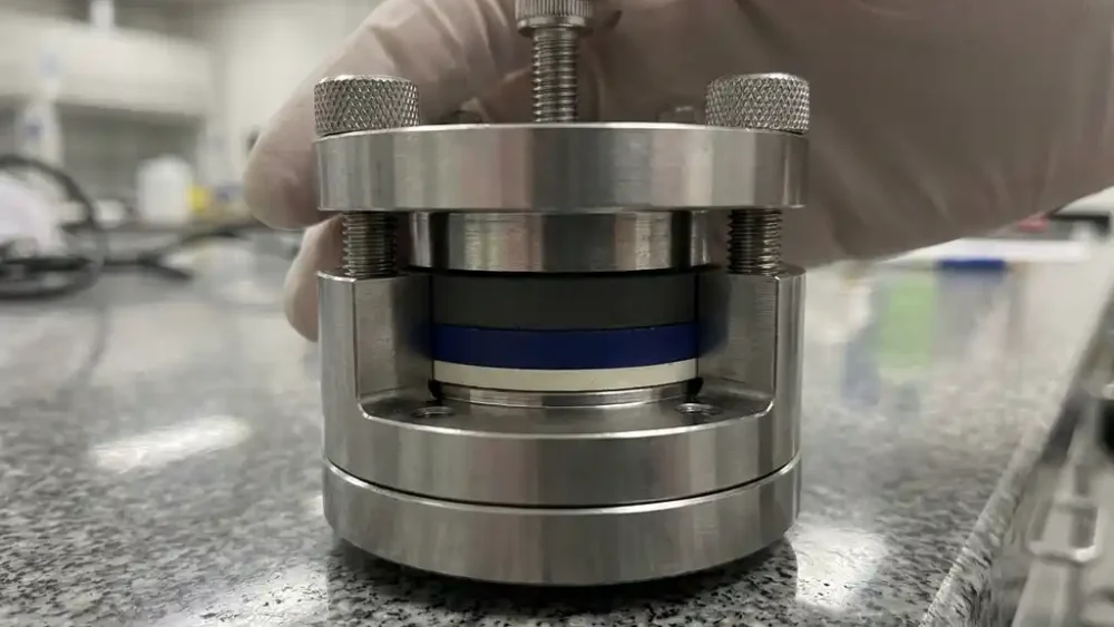 Stainless steel solid-state battery test cell compressed on a granite lab bench, revealing ceramic electrolyte and electrode layer cross-sections.