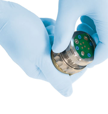 Two blue-gloved hands hold and inspect a small metallic medical device with a green circuit-like top.