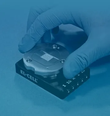 Gloved hand placing a round metal cell into a black fixture labeled EL-CELL in a blue-tinted lab setup.